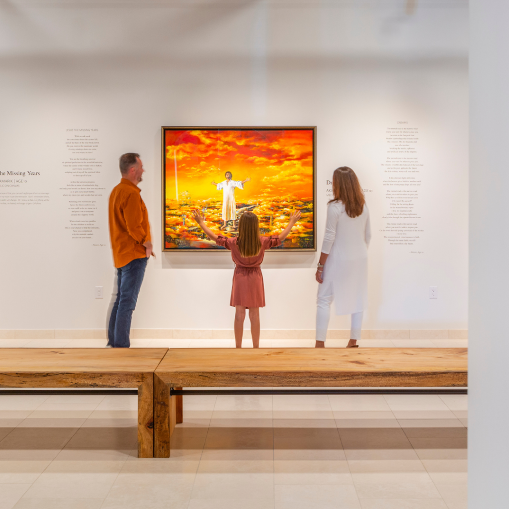 A family of three viewing Akiane Kramarik's painting "Jesus: The Missing Years" in a gallery setting. The young girl in the center reaches her arms outward, mimicking the pose of Jesus in the painting, which depicts Him in a white robe hovering over a cityscape of past, present, and future architecture, illuminated by a vivid orange sunset sky.