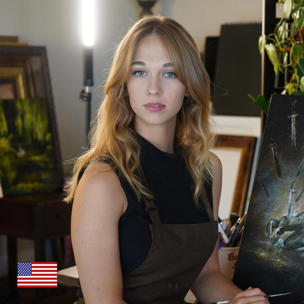 A young woman with long blonde hair in a black top and brown apron sits in an art studio, holding a paintbrush. Paintings and supplies are visible in the background. A small U.S. flag icon is in the lower left corner.