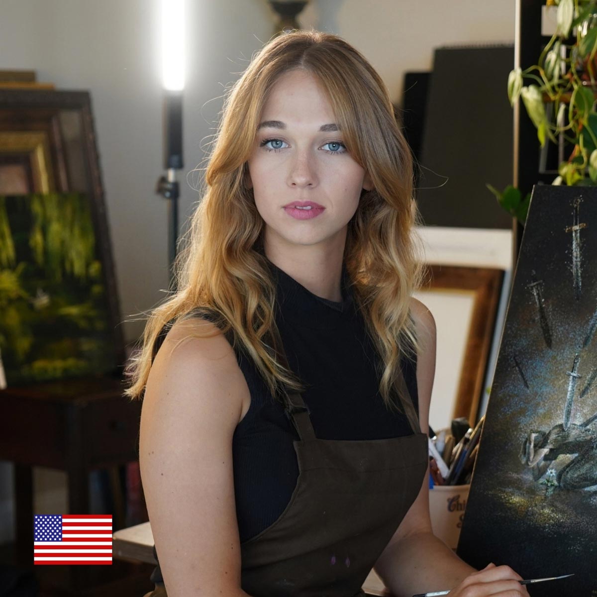 A young woman with long blonde hair in a black top and brown apron sits in an art studio, holding a paintbrush. Paintings and supplies are visible in the background. A small U.S. flag icon is in the lower left corner.