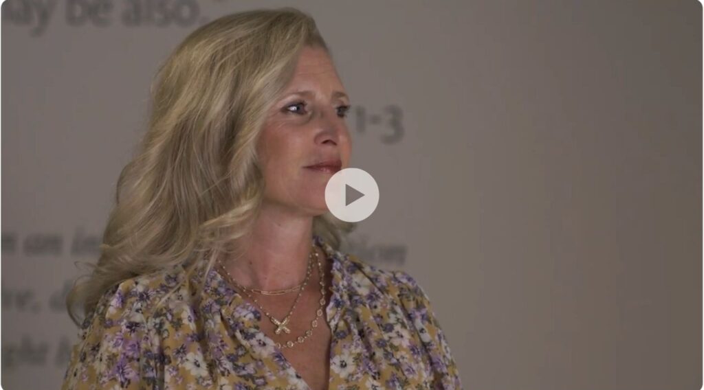 A woman with long blonde hair wearing a floral blouse and layered necklaces stands in front of a blurred background with indistinct text. She is looking slightly to the side and appears thoughtful.