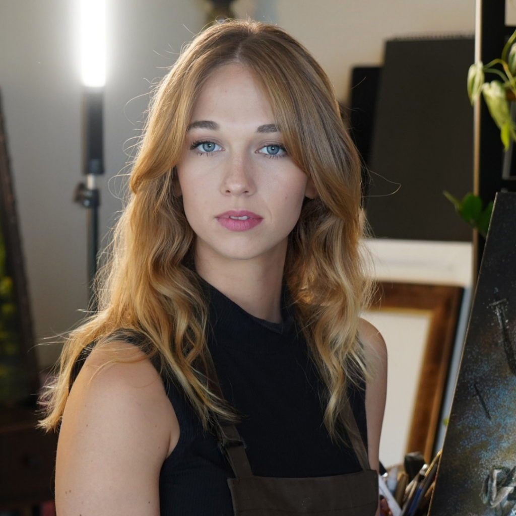 Master Fine Artist Akiane Kramarik with long blonde hair and blue eyes stands indoors, wearing a dark sleeveless top and apron, with an easel and painting supplies in the background.