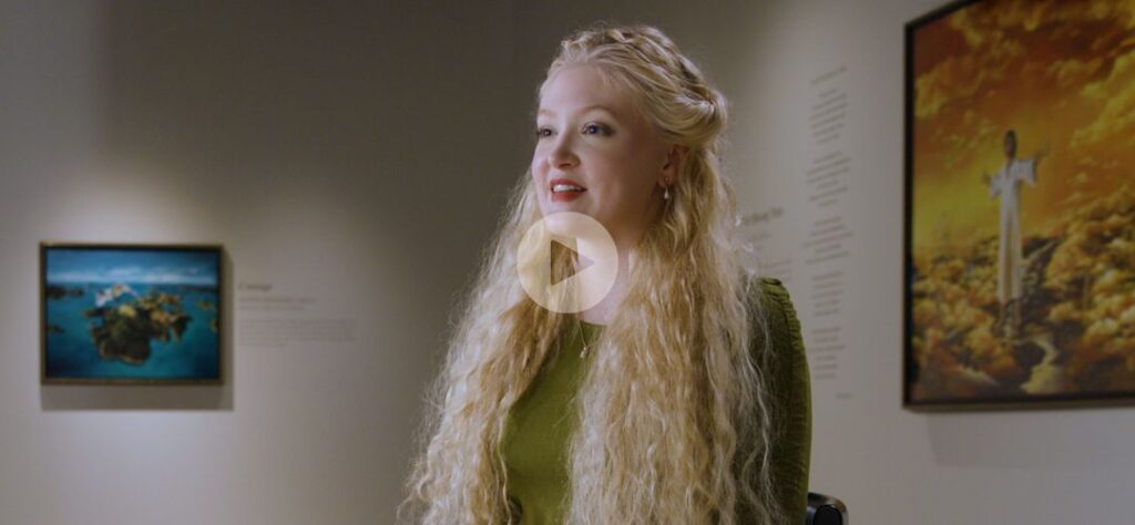 A woman with long, wavy blonde hair sits in an art gallery, speaking. She wears a green top and is surrounded by paintings on the walls. A play button icon appears over the center of the image.
