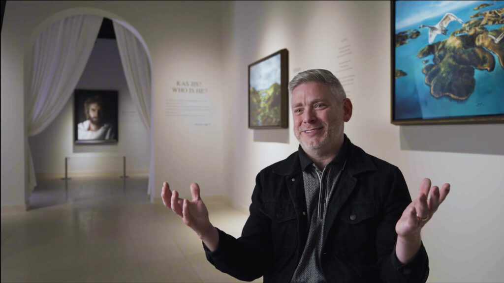 A man with gray hair and a beard, wearing a black jacket, sits smiling and gesturing with open hands in an art gallery with paintings and white walls in the background.