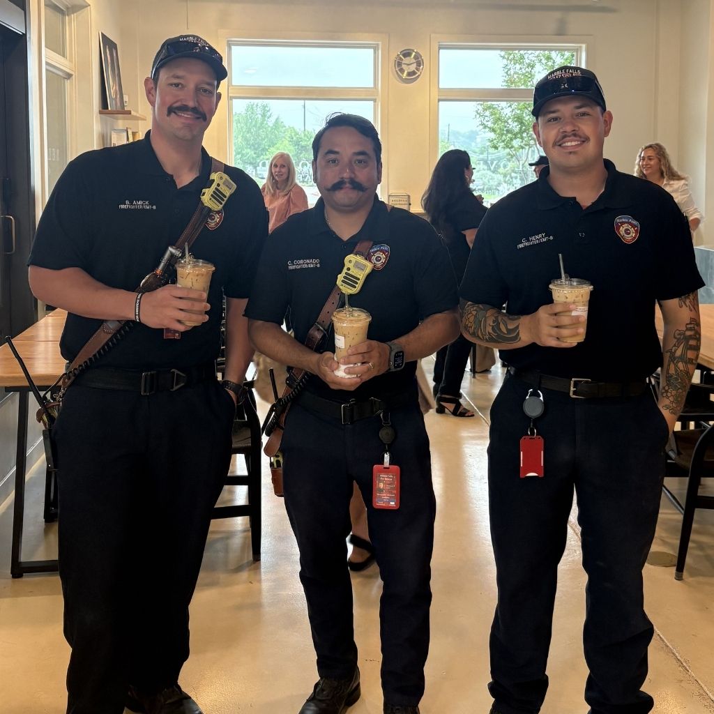 Three firefighters in navy uniforms pose with iced coffees inside a bright cafe area.