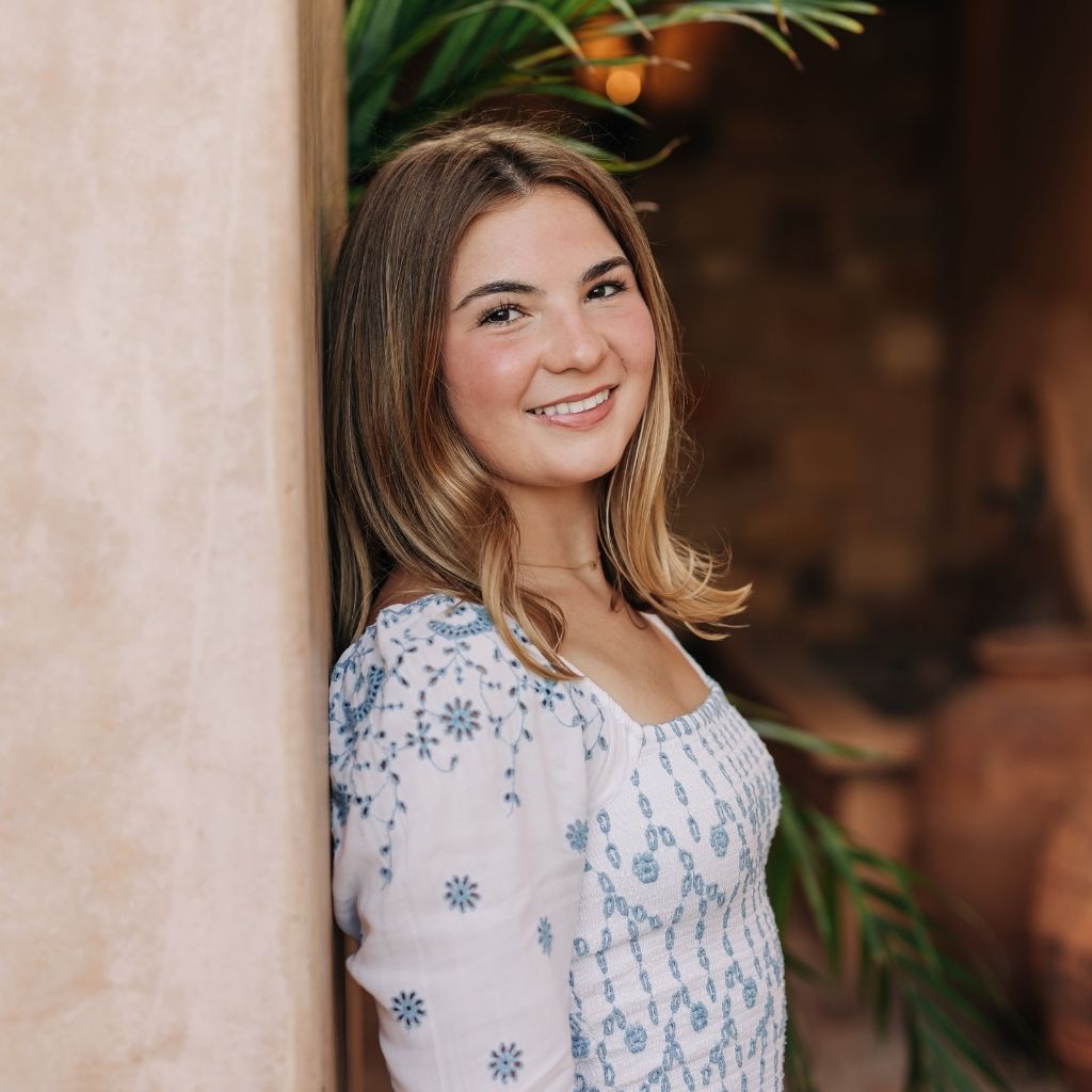 Smiling woman leaning against a stone pillar outdoors, wearing a white embroidered blouse.