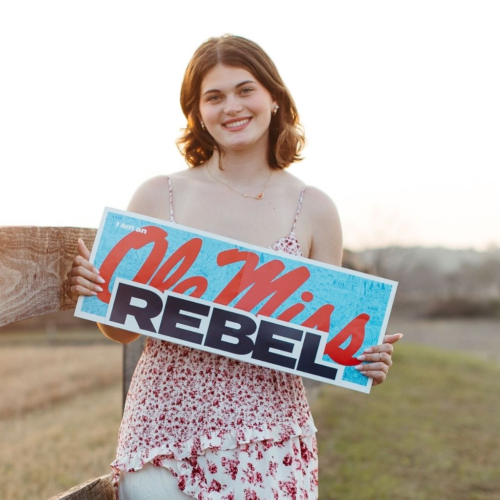 Smiling young woman in a floral dress outdoors, holding a blue sign that reads 'REBEL'.