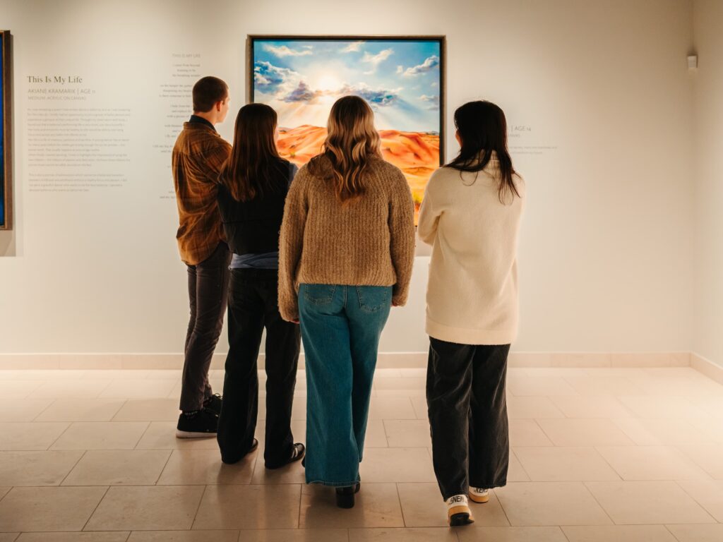 Four people stand with backs to the camera, viewing a bright desert landscape painting in a white-walled gallery.