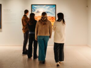 Four people stand with backs to the camera, viewing a bright desert landscape painting in a white-walled gallery.