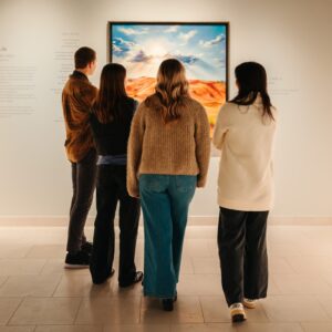 Four people stand with backs to the camera, viewing a bright desert landscape painting in a white-walled gallery.
