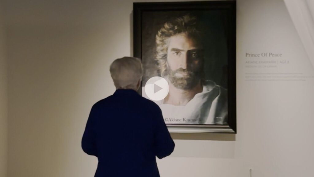 Older person with gray hair in a dark blue jacket stands with their back to the camera, viewing a large framed portrait in a museum gallery (titled 'Prince Of Peace').