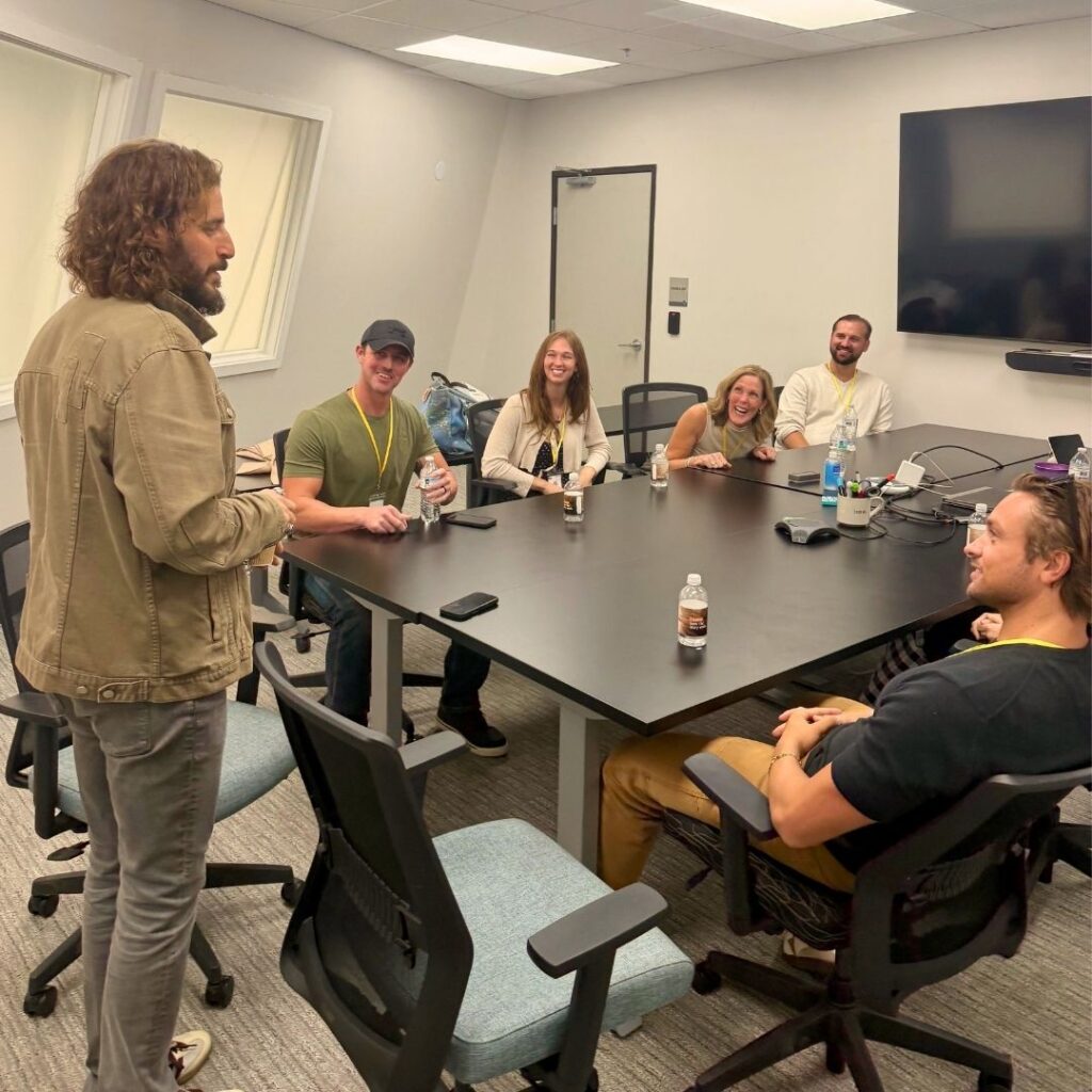 A man stands and speaks to six people sitting around a conference table in a meeting room. Everyone appears engaged and smiling. Drinks and bottles are on the table, and a large TV is mounted on the wall.