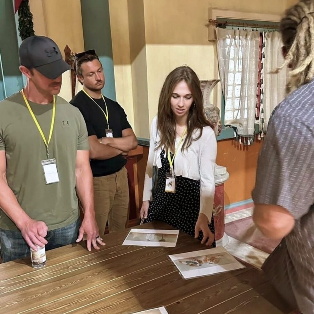 Four people stand around a wooden table looking at printed papers. One woman gestures toward the papers while the others watch. They all wear lanyards and are in a room with warm lighting and decorative walls.