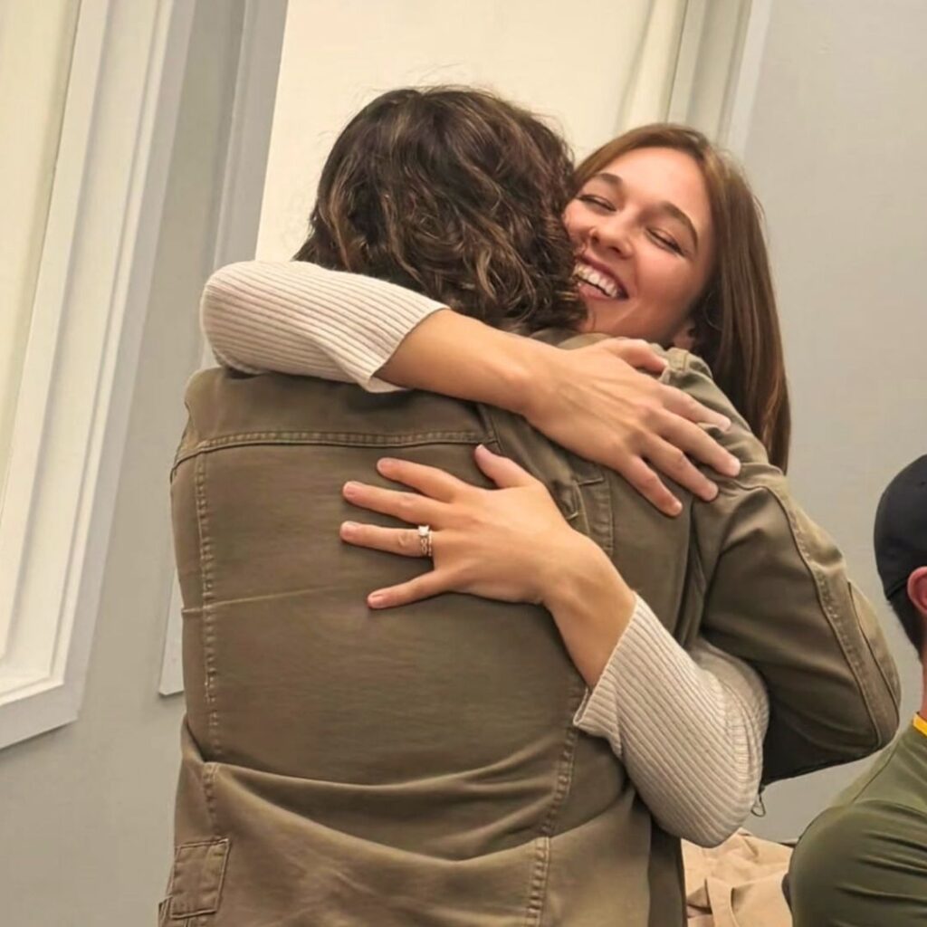 Two women are hugging each other warmly. One woman is smiling joyfully with her eyes closed, and a ring is visible on her finger. The setting appears to be indoors, with light-colored walls in the background.