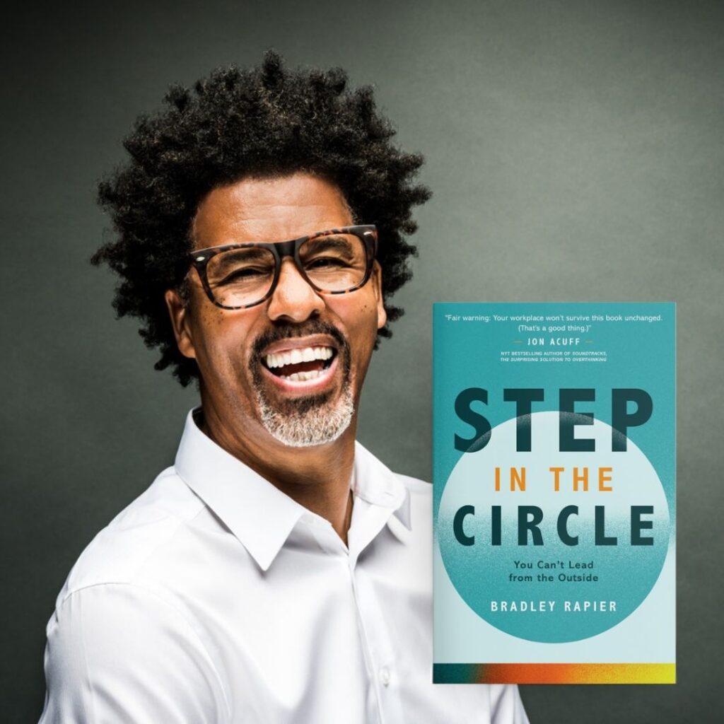 A smiling man with curly hair, glasses, and a beard, wearing a white shirt, stands next to the book Step in the Circle: You Cant Lead from the Outside by Bradley Rapier against a gray background.