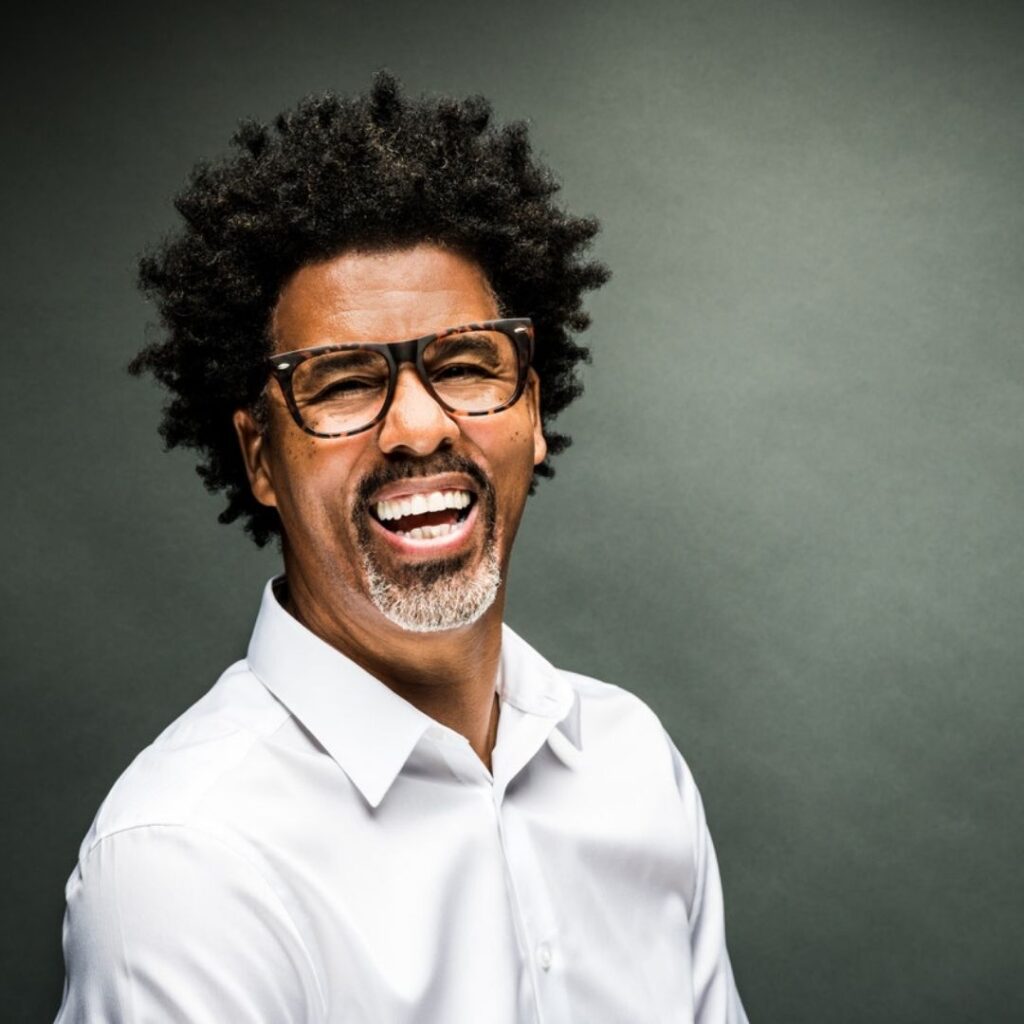 A man with curly hair, glasses, and a short beard smiles widely while wearing a white shirt against a plain dark background.