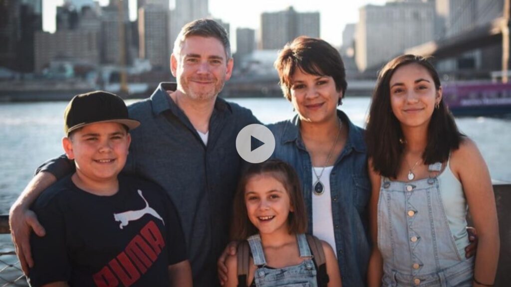 A smiling family of five poses outdoors near a river, with a city skyline and bridge in the background. The group includes two adults and three children, all standing close together in casual clothing on a sunny day.