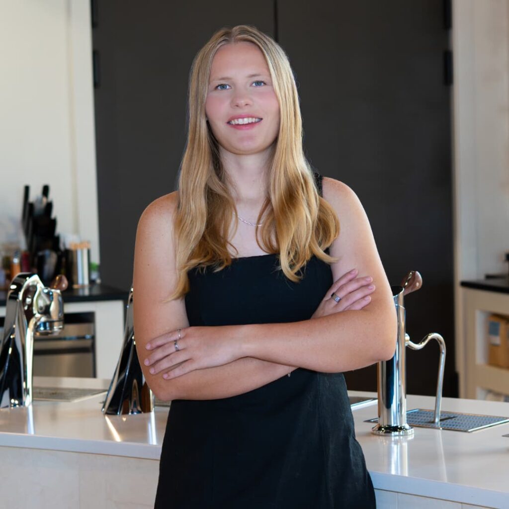 Smiling young woman with long blonde hair and a black sleeveless top standing with arms crossed behind a white bar counter in a modern kitchen setting.