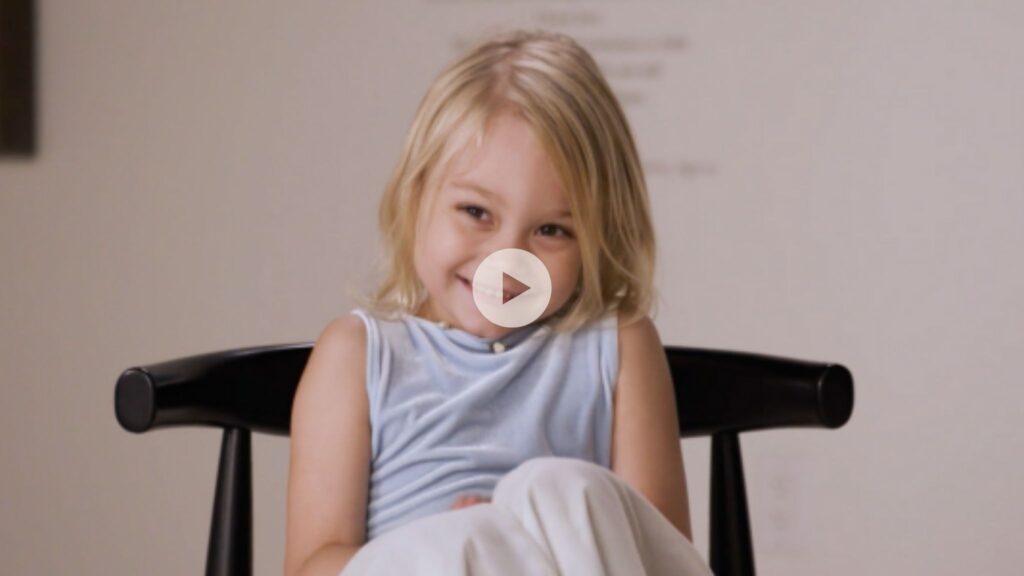 A young child with blonde hair smiles shyly while sitting on a black chair, wearing a light blue sleeveless top. The image has a play button overlay, indicating it is a video thumbnail.
