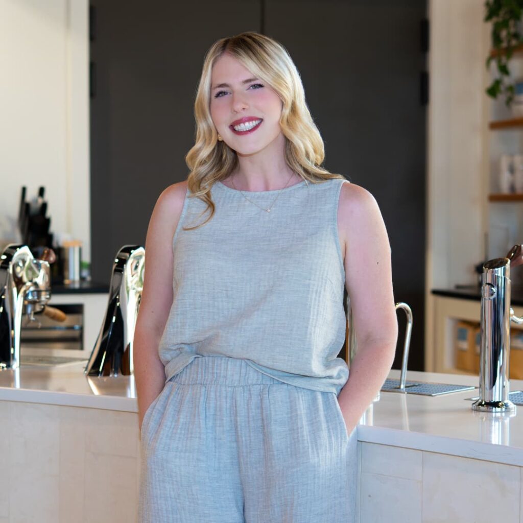 Smiling woman in a light blue sleeveless jumpsuit stands at a white cafe counter.