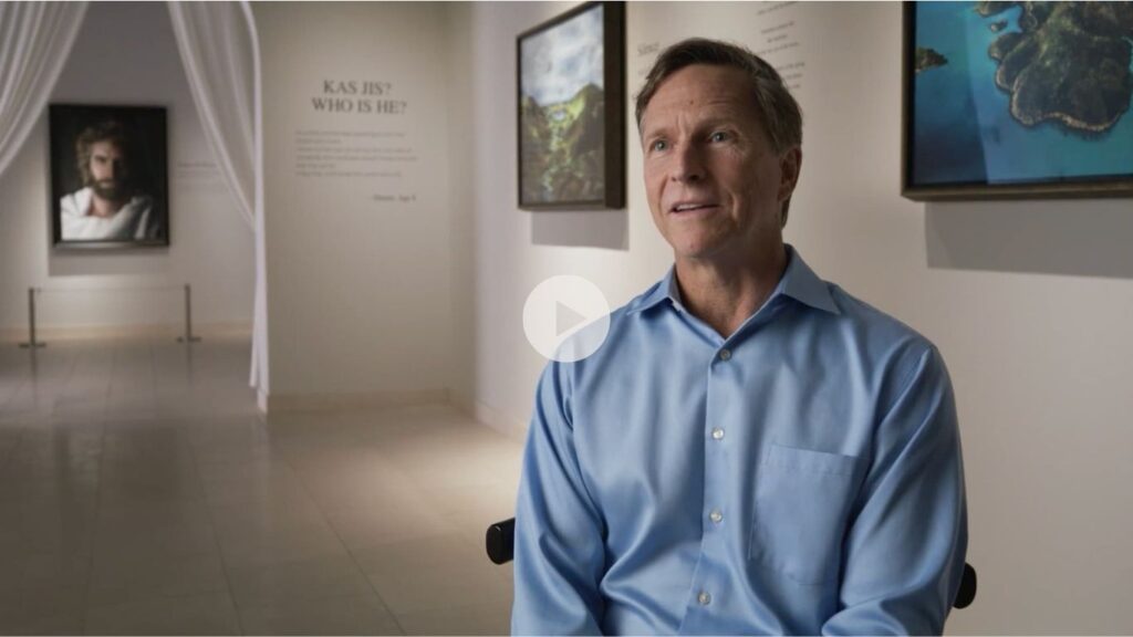 A man in a light blue shirt sits and speaks in an art gallery with paintings and a wall text reading “KAS HIS? WHO IS HE?” displayed behind him. Soft lighting highlights the gallerys calm atmosphere.