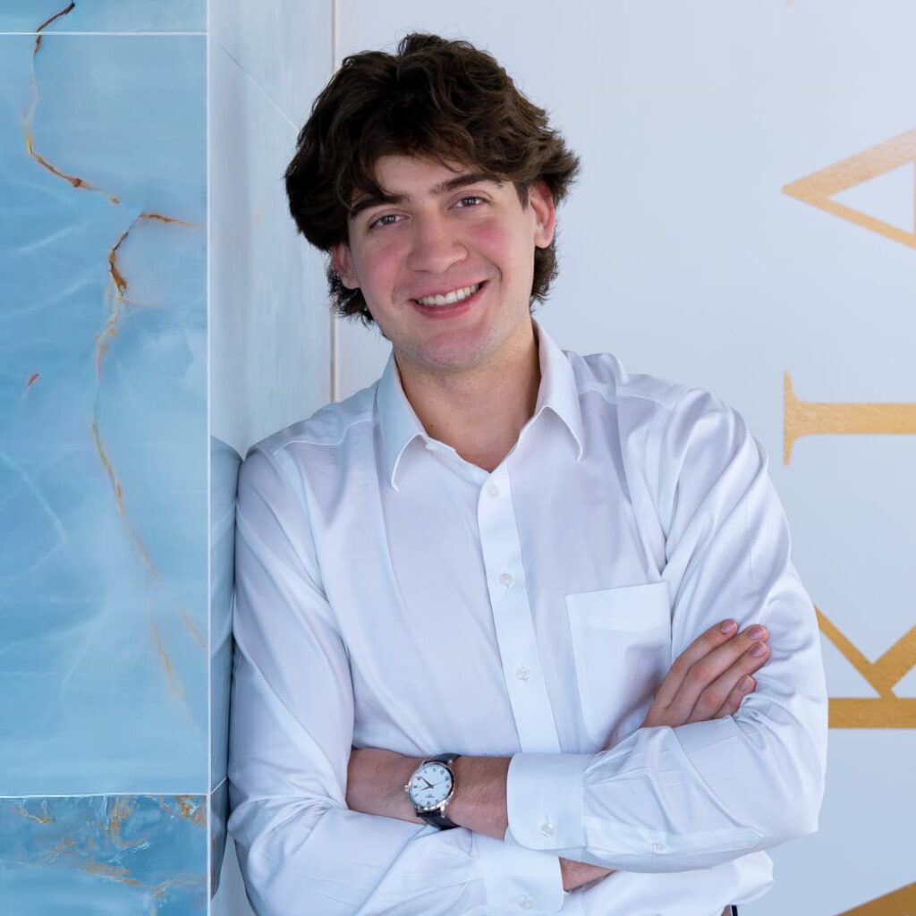 Young man in a white shirt leaning against a turquoise marble wall, arms crossed and smiling.