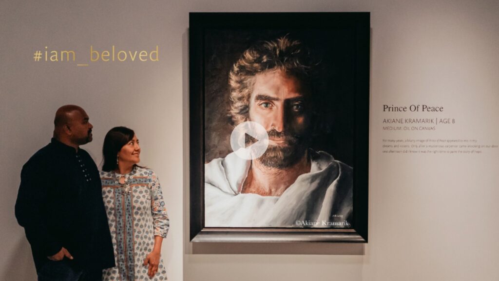 A man and woman stand next to a large painting of Jesus titled Prince of Peace by Akiane Kramarik, age 8, displayed on a gallery wall. The text #iam_beloved appears in the top left corner.