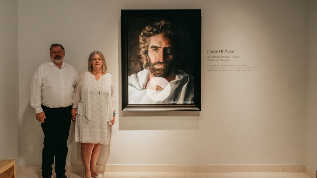 A man and a woman stand side by side in front of a framed portrait of Jesus titled Prince of Peace, displayed on a white wall with a description next to it. Both are dressed in white.
