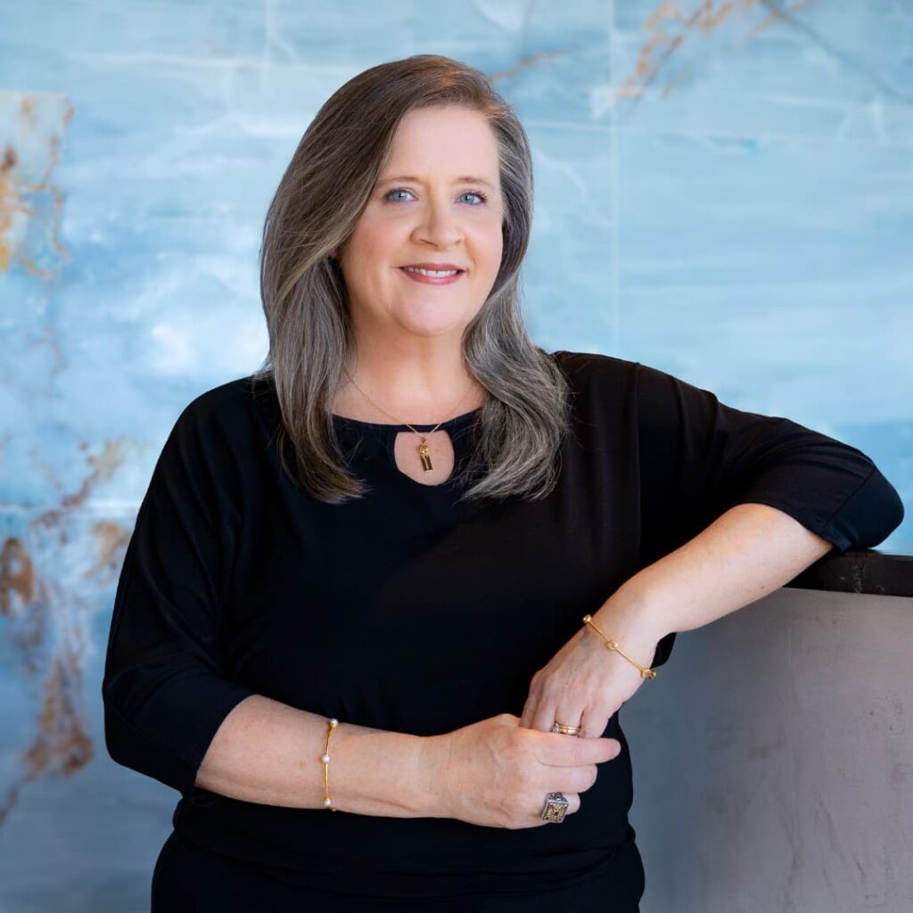 Smiling woman with gray hair in a black top, leaning on a railing against a blue wall.