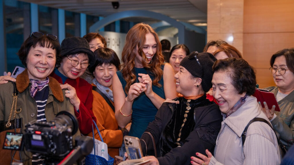 A group of women smiling and laughing together indoors, with one woman in a green dress at the center. Some women are holding phones and cameras, capturing the joyful moment.