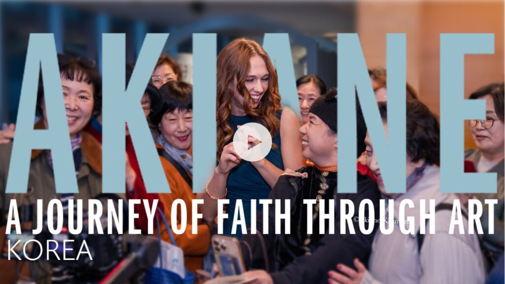 A smiling woman stands among a group of excited people taking photos. Large text reads: “AKIANE: A Journey of Faith Through Art, Korea.” A play button is over the woman, suggesting a video.