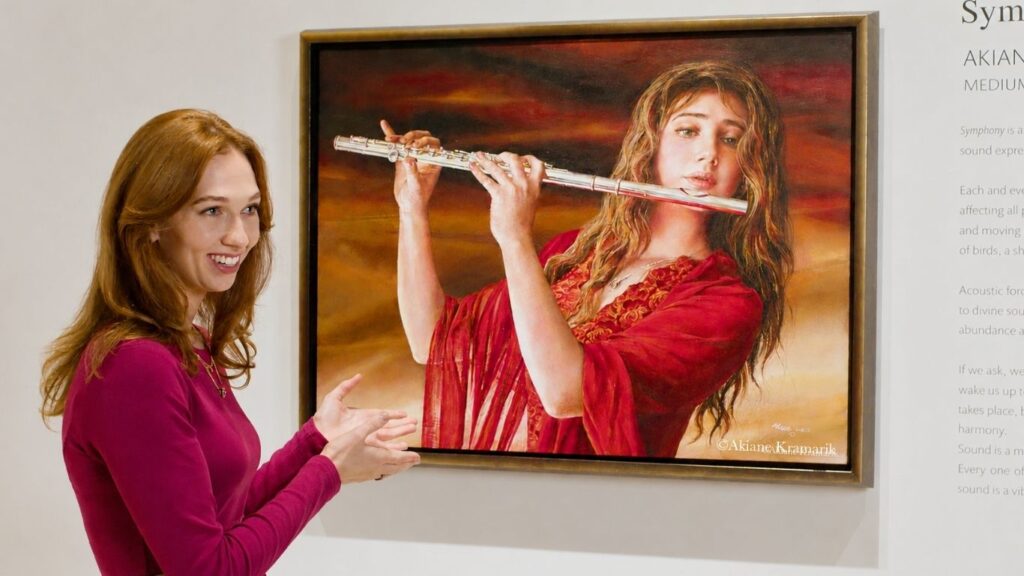 A smiling woman with long red hair and a pink top gestures toward a framed painting of a woman in red playing a flute, displayed on a white wall.