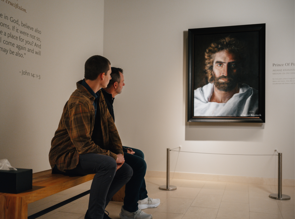 Two men in casual jackets sit on a wooden bench, looking at a large framed portrait of a bearded man in a museum setting.