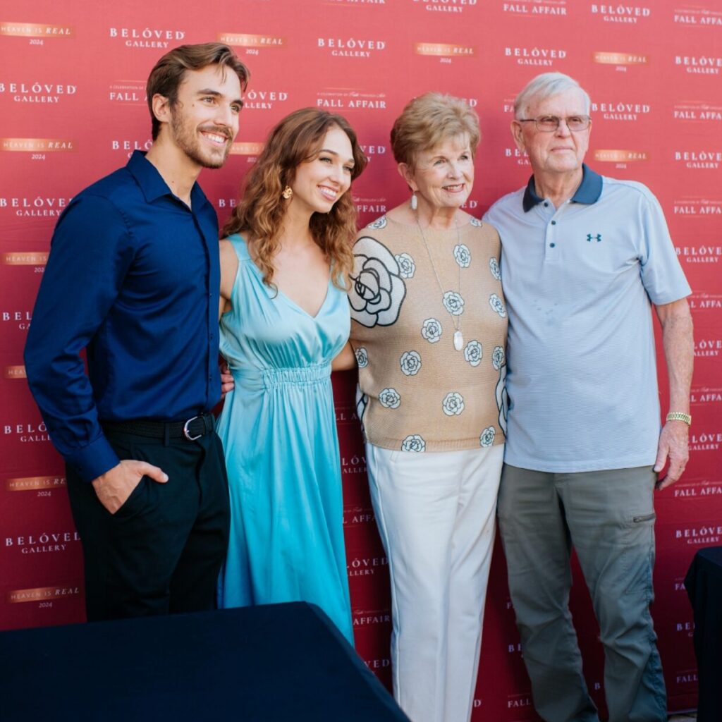 Four people stand smiling in front of a red step-and-repeat backdrop. Two are younger adults dressed formally, and two are older adults in casual clothes. They pose closely together outdoors.