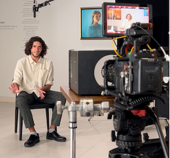 A man sits on a chair being filmed in a studio, gesturing with his hands. Professional camera equipment and a microphone are visible. Portraits hang on the wall in the background.