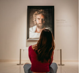 A woman with long hair, wearing a red off-shoulder top and jeans, sits on the floor in an art gallery, facing and looking at a framed portrait of a bearded man on the wall.