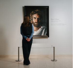 A person with long hair, seen from behind, stands in a museum looking at a framed portrait of Jesus titled Prince of Peace by Akiane Kramarik, with a description on the wall beside it.