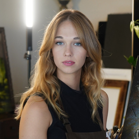 A young woman with long, wavy blonde hair and blue eyes stands indoors, wearing a dark top and apron, looking directly at the camera with a neutral expression. Art supplies and paintings are visible in the background.