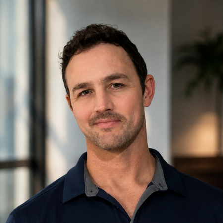 A man with short dark hair and trimmed facial hair, wearing a navy collared shirt, stands indoors with a soft smile. The background is softly lit with a window and a blurred plant visible.