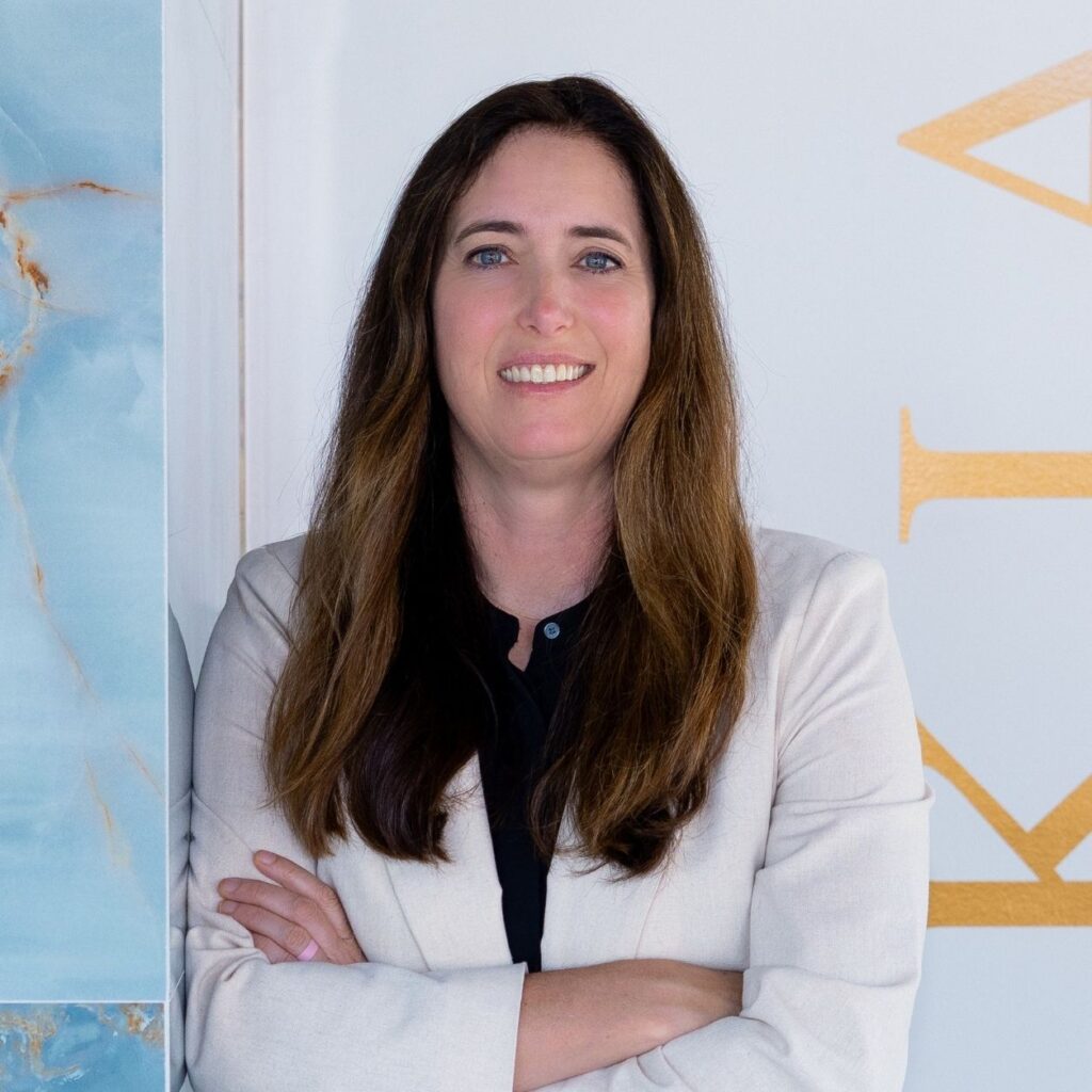 A woman with long brown hair wearing a light blazer and black shirt stands smiling with arms crossed in front of a white and blue marble wall.