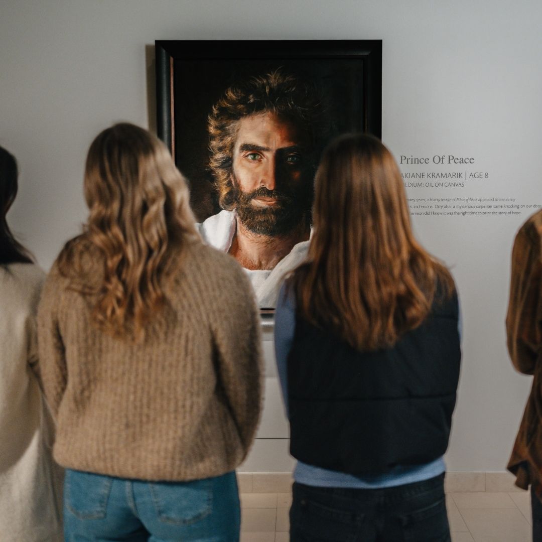 Four people stand closely together, facing and observing a framed portrait titled Prince of Peace, which depicts a man with long hair and a beard, displayed on a white gallery wall.