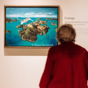 A person in a red coat views a framed painting of islands and a white bird flying over blue water, displayed in an art gallery.