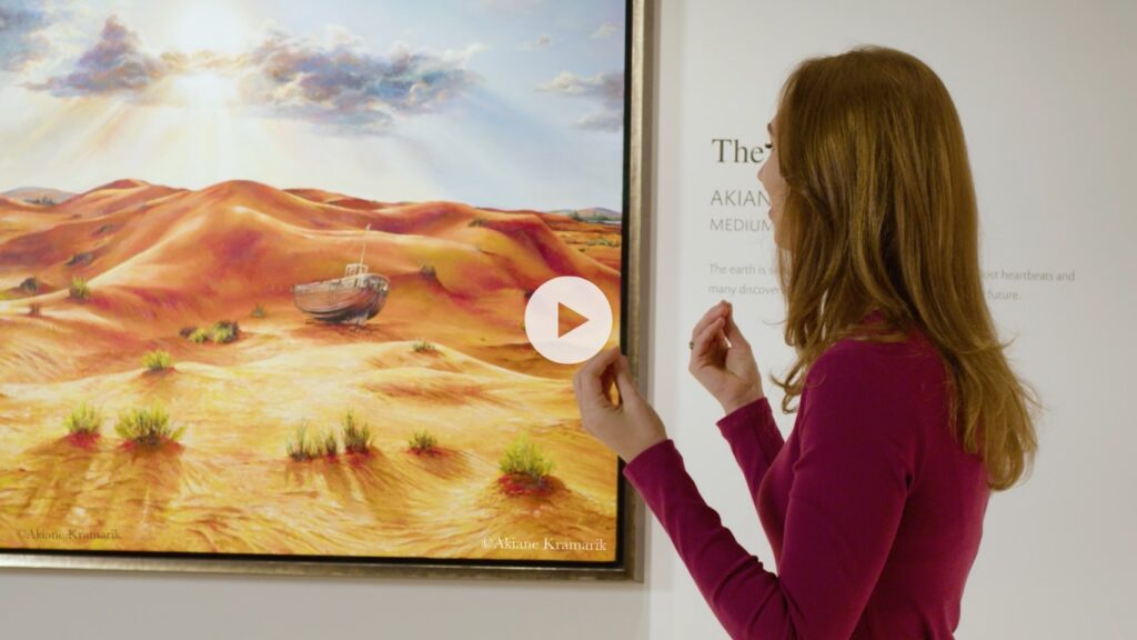 A woman in a magenta top gestures while viewing a vibrant painting of a desert landscape with an abandoned boat, displayed in an art gallery.