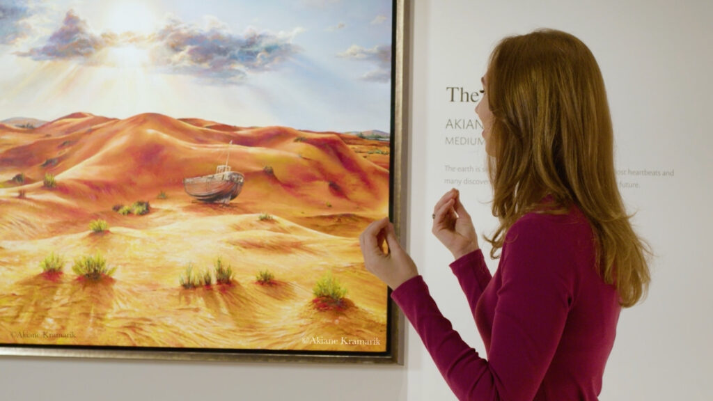 A woman with brown hair in a magenta top gestures while viewing a framed painting of a boat on sandy, red-orange desert dunes in an art gallery.