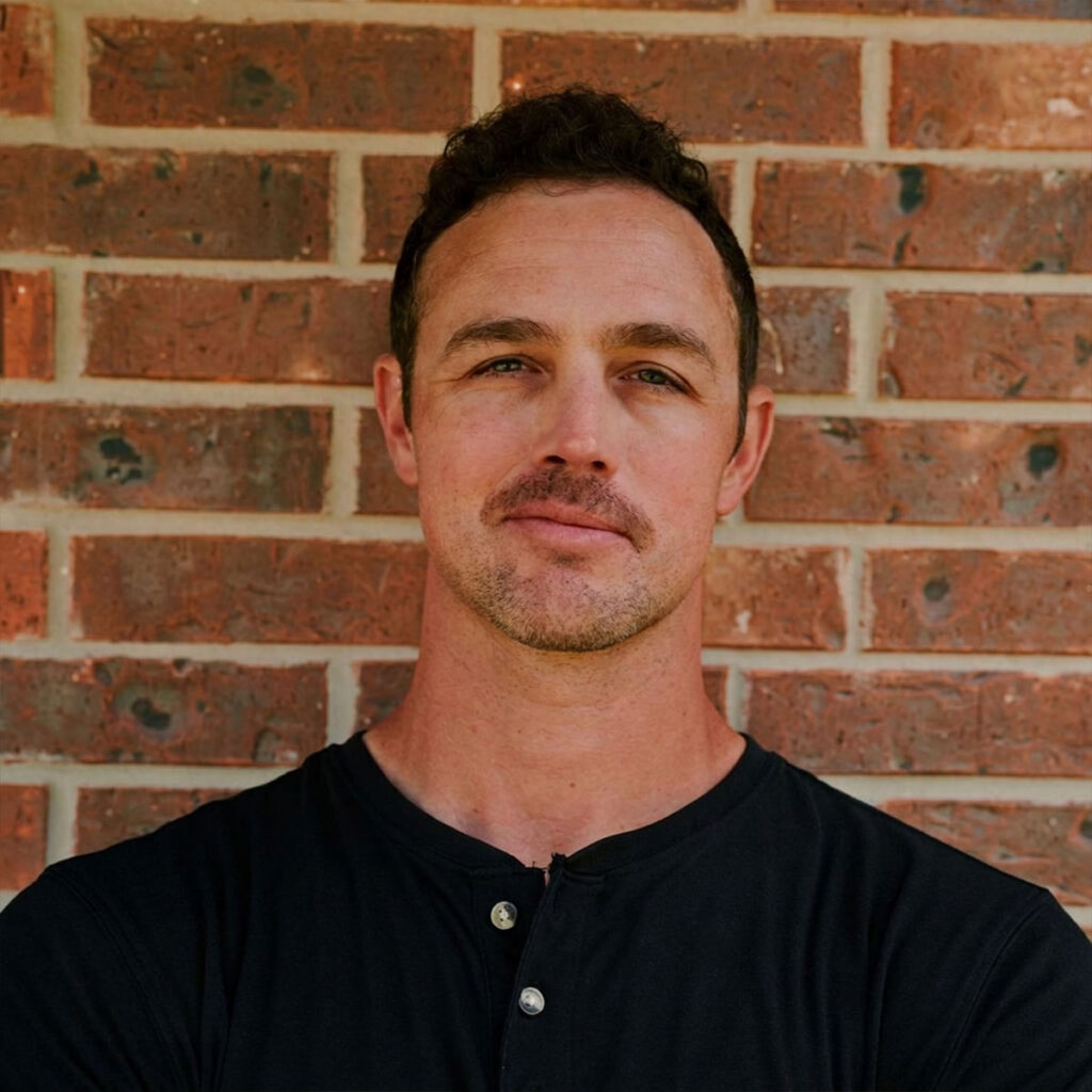 A man with short, curly dark hair and light stubble stands in front of a brick wall, wearing a black henley shirt and looking directly at the camera with a neutral expression.