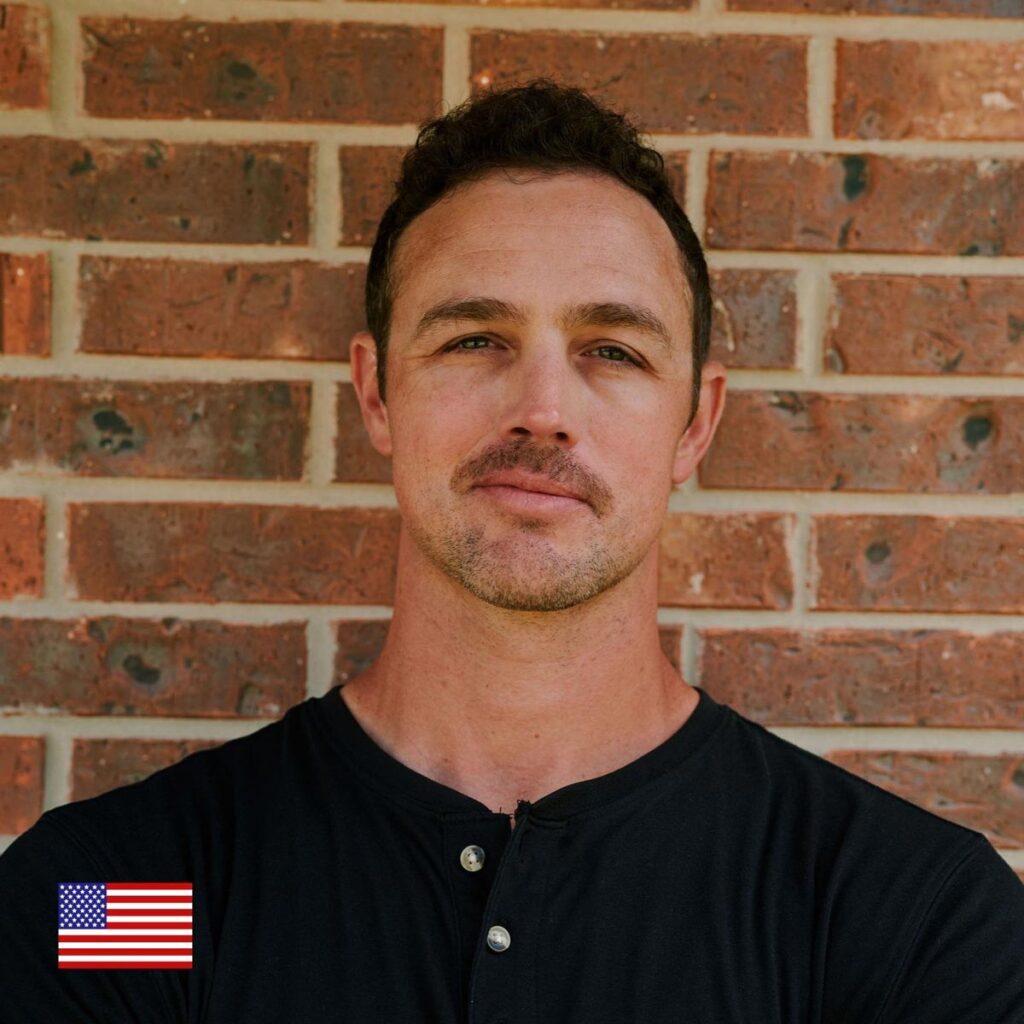 A man with short brown hair and a trimmed beard stands in front of a brick wall, wearing a black henley shirt. A small American flag icon appears in the bottom left corner of the image.