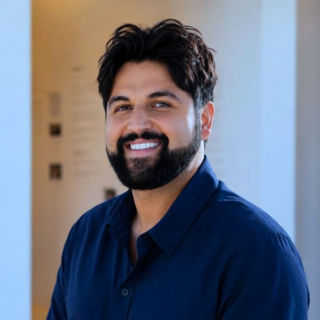 A man with dark hair and a beard, wearing a dark blue shirt, smiles while standing indoors in a well-lit room with light-colored walls.