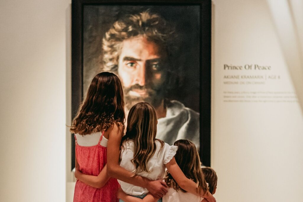 Three girls stand close together, arms around each other, looking at a large framed portrait of a bearded man in a gallery labeled Prince Of Peace..
