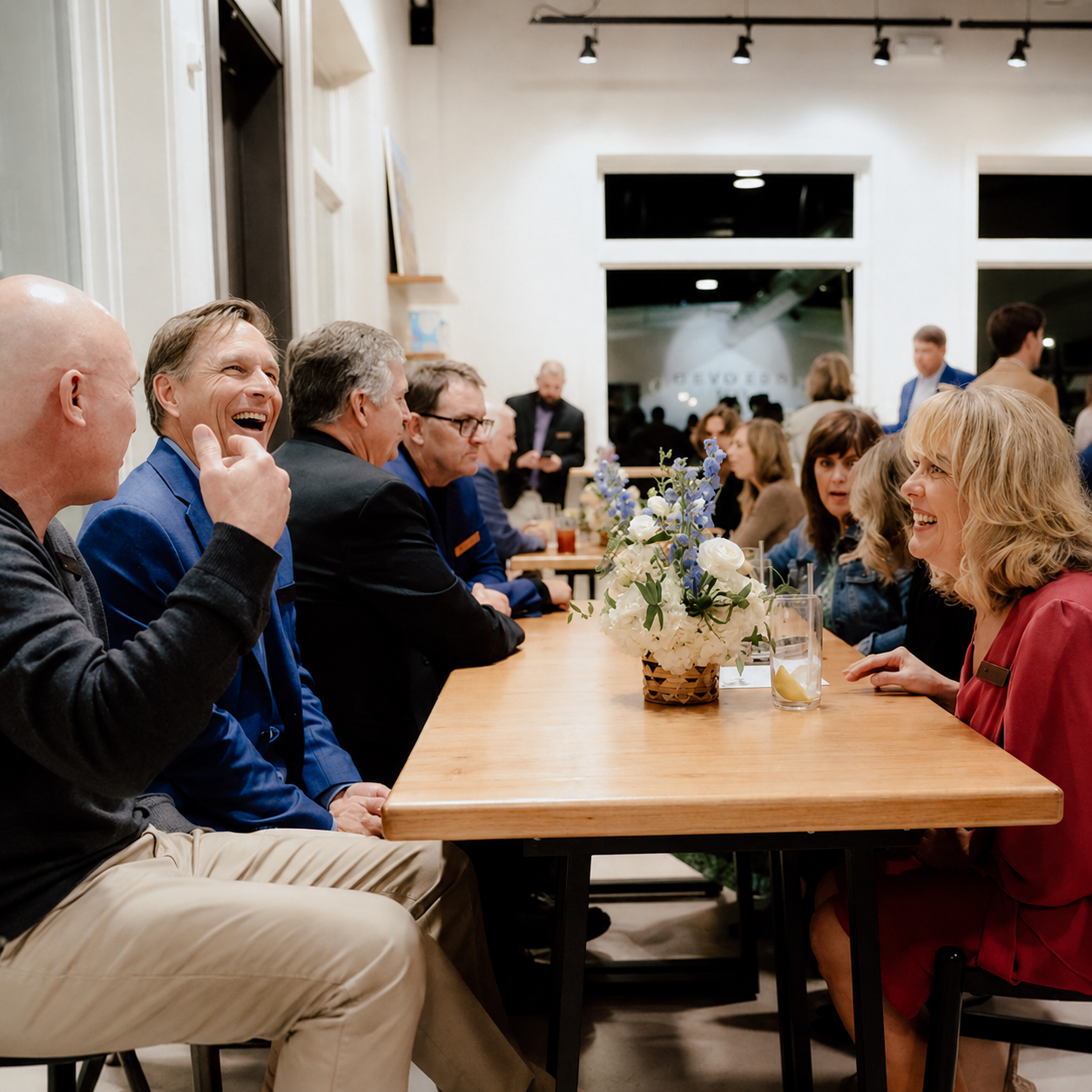 People sitting at a long wooden table chatting and laughing at a social gathering, with a flower centerpiece in the middle.