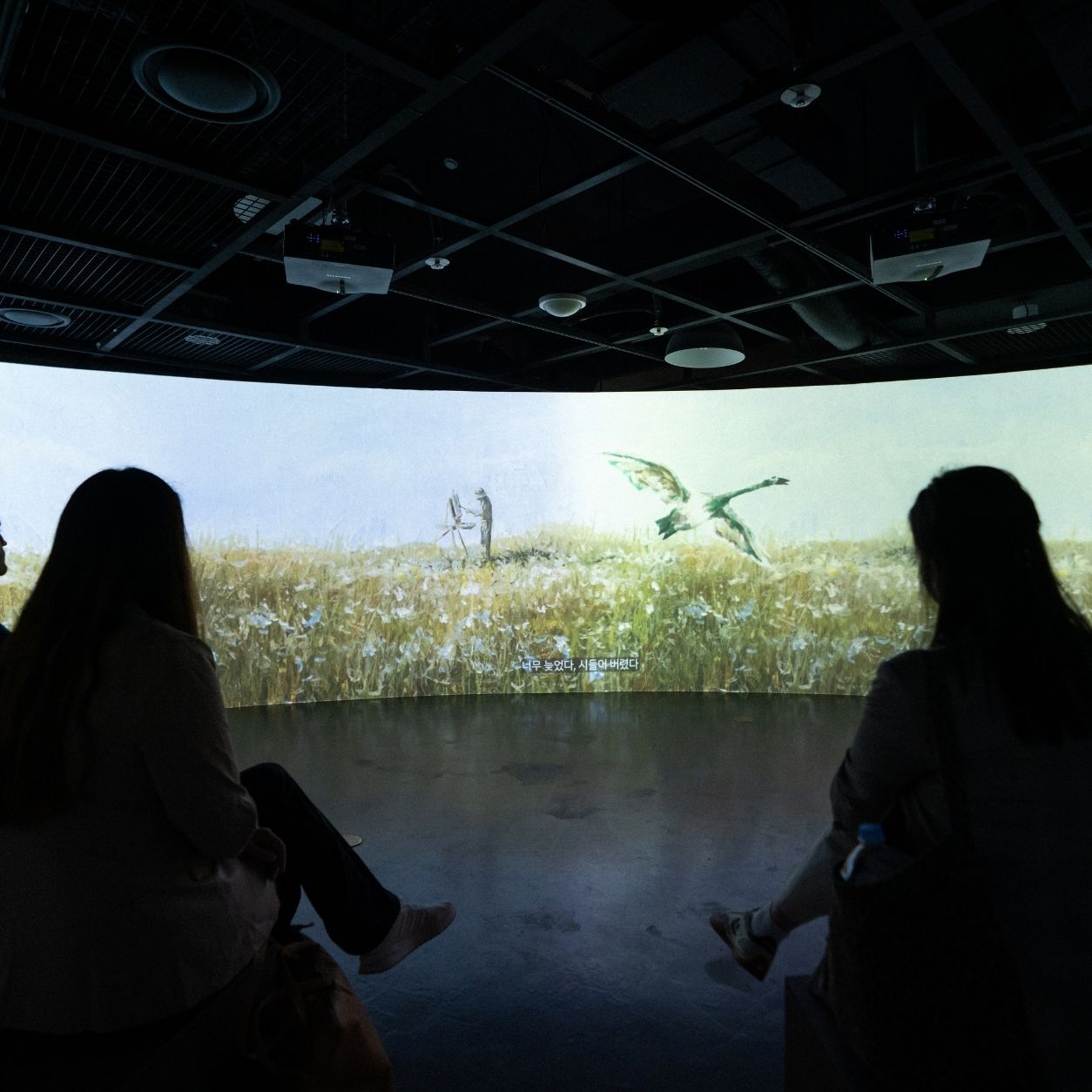 Three people sit in a dark room watching a panoramic screen showing a field with a large bird or dragon and a person standing in the grass.