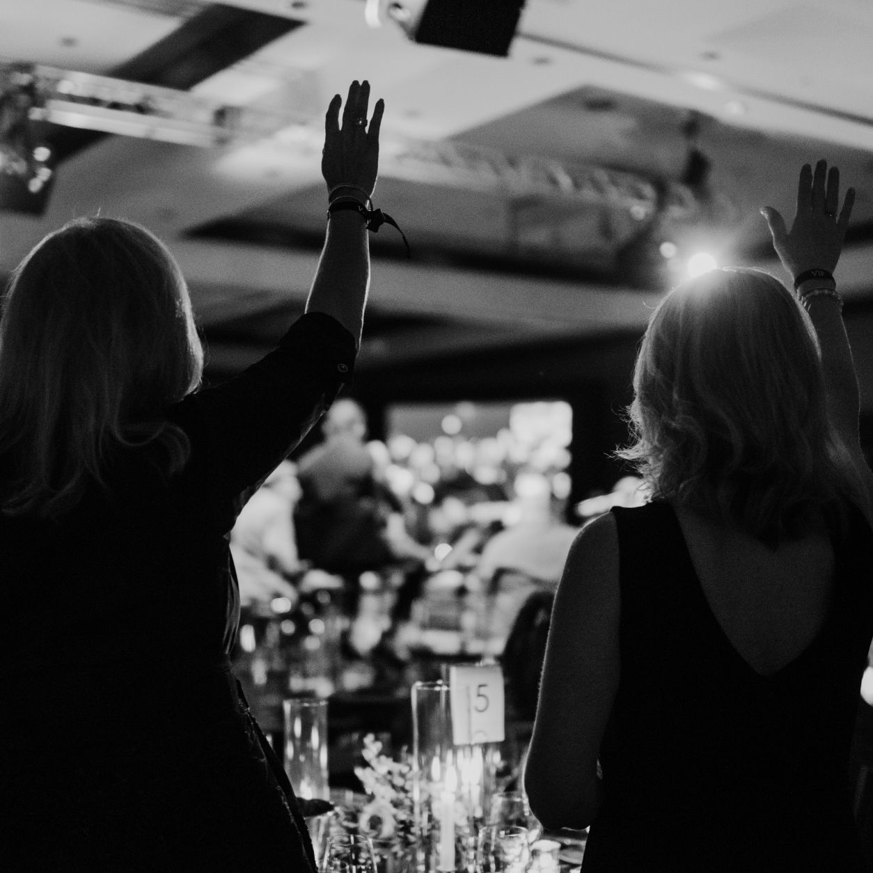 Two people stand with arms raised at an indoor event, facing a stage in a dimly lit room filled with seated guests. The image is in black and white, creating a festive and energetic atmosphere.